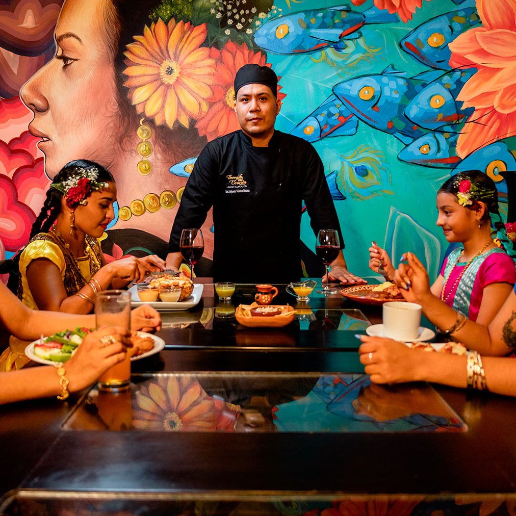 Nuestro chef junto a la mesa, frente al mural de la tehuana, con comensales en traje regional